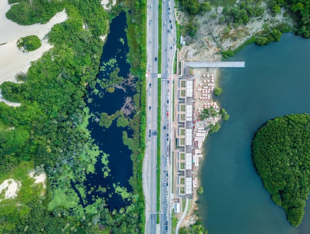 Vista aérea de uma faixa litorânea urbanizada, com estrada, áreas verdes e construções próximas à costa.